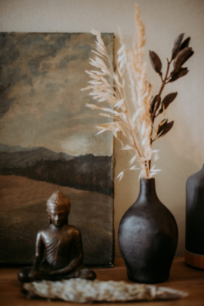 Bronze-Buddha-Statue neben einer dunklen Vase mit getrocknetem Pampasgras und einem abstrakten Landschaftsbild – ruhige und natürliche Dekoration im Behandlungsraum.