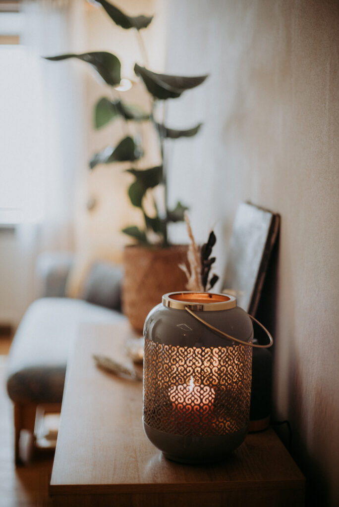 Graue Kerzenlaterne mit warmem Kerzenlicht auf einem Sideboard, im Hintergrund eine große Zimmerpflanze – stimmungsvolle Beleuchtung im Entspannungsbereich der Massagepraxis.